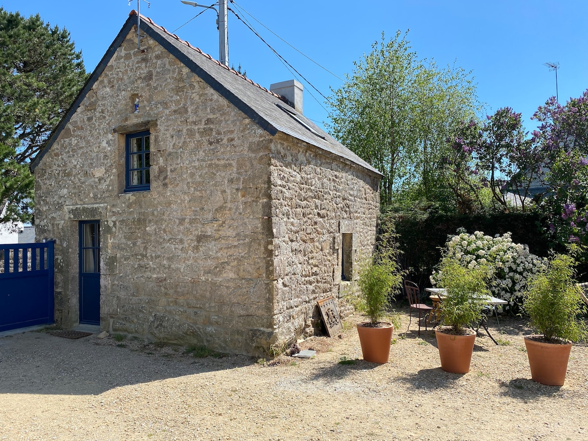 A quaint stone cottage is visible, featuring a blue door and window framing charming garden views. Potted plants adorn the exterior, while a table and chairs are arranged outside, inviting relaxation. Lush greenery surrounds the area, suggesting a tranquil outdoor setting.