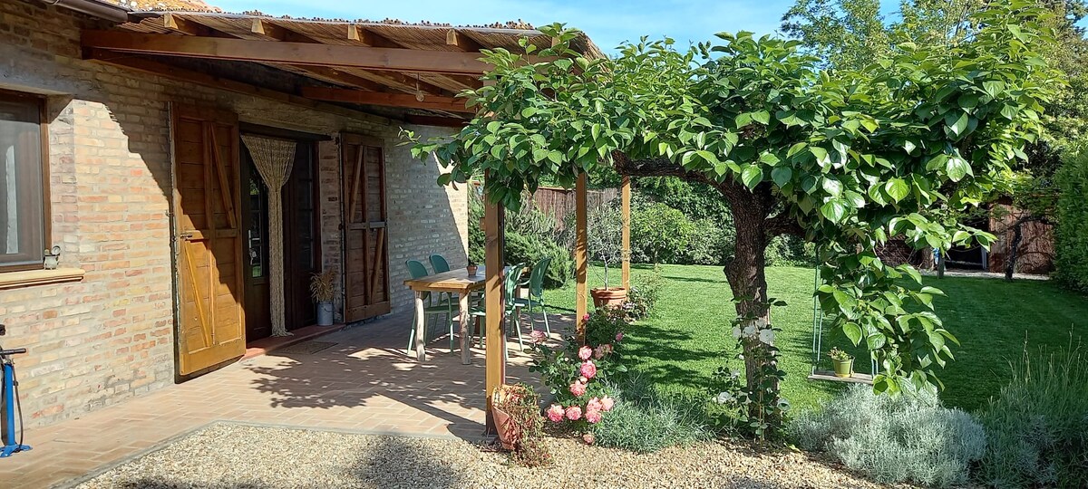 A charming patio area is featured, with wooden beams overhead providing shade. Green chairs and tables are arranged for outdoor seating, surrounded by lush greenery and blooming flowers. The well-kept garden invites relaxation, with a spacious lawn stretching beyond.