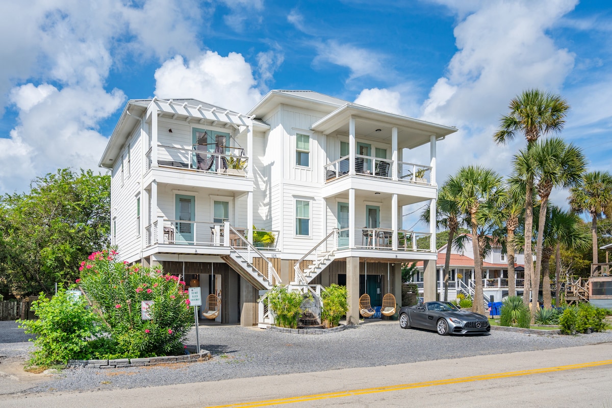 A modern three-story duplex is displayed under a clear blue sky, featuring a light-colored exterior. Palm trees and vibrant greenery surround the property. The front porch includes two hanging chairs, providing additional outdoor seating. A spacious driveway accommodates a parked vehicle.