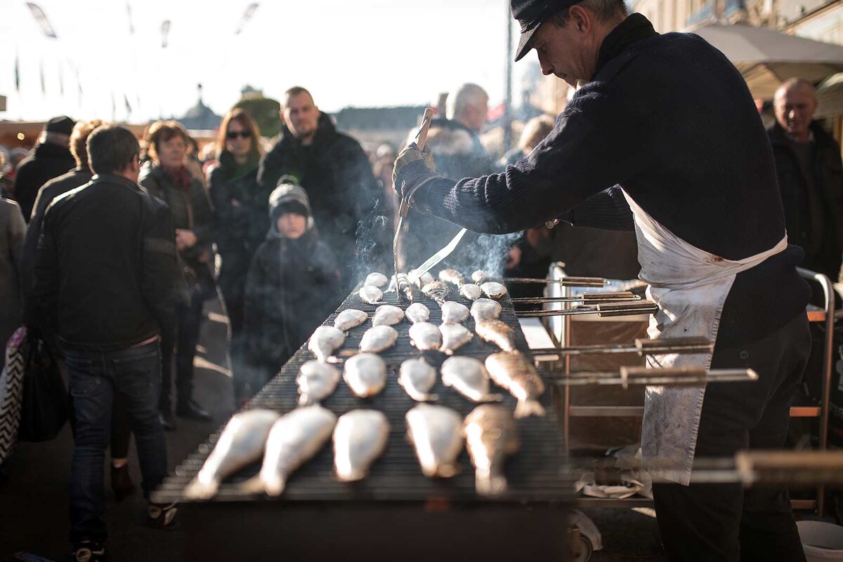 A bustling outdoor market scene is presented with a vendor grilling fish over an open flame. Smoke rises gently, while a crowd of shoppers observes, visible in the background. The griddle is lined with neatly arranged fish, showing the lively atmosphere of the market.