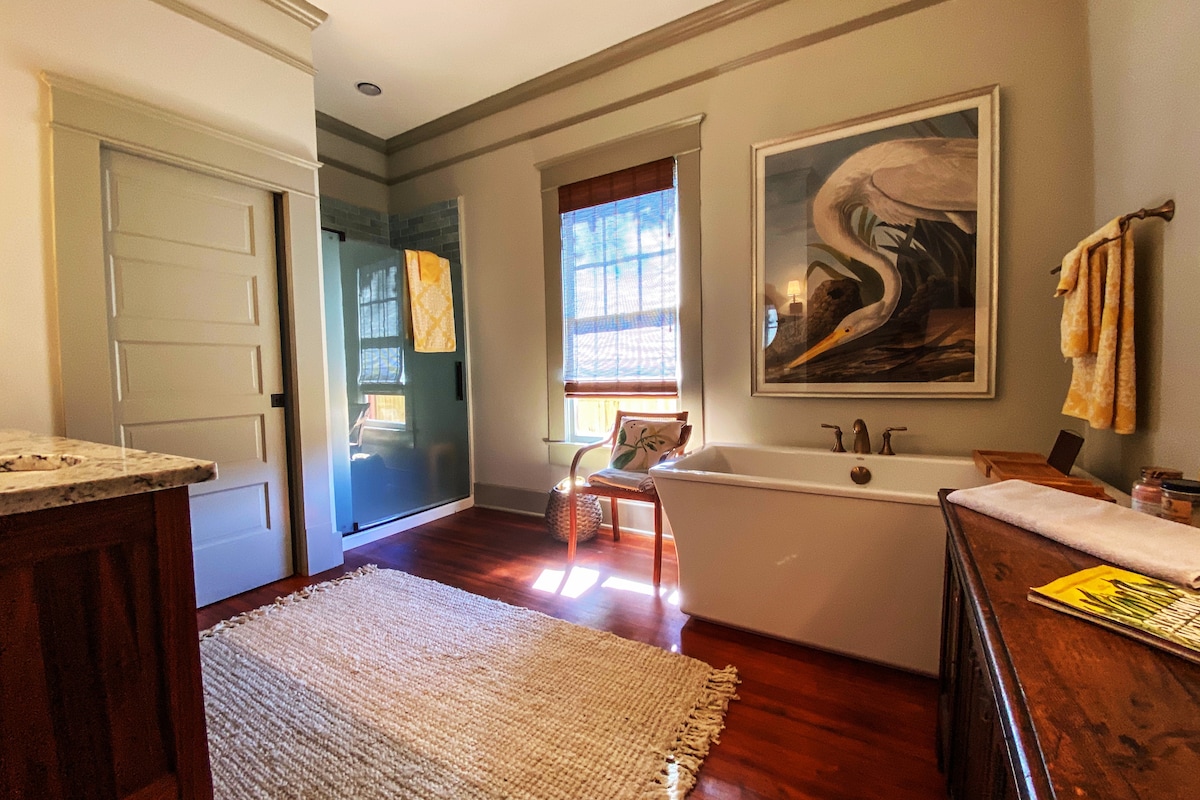 The bathroom features a soaking tub positioned beside a wooden console. A glass-enclosed shower is visible, with natural light filtering through the window. A piece of art adorns the wall, and a textured area rug covers the hardwood floor, creating a serene atmosphere.