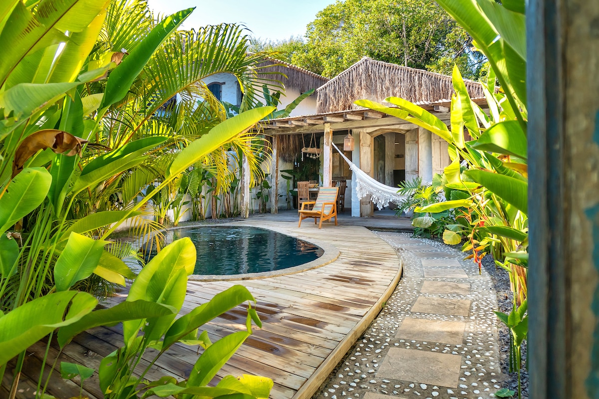 A private pool is surrounded by a wooden deck and lush tropical vegetation. A hammock is visible, draped between posts, with a wooden lounge chair positioned nearby. Natural stone pathways lead to the entrance of the house, creating an inviting outdoor space.