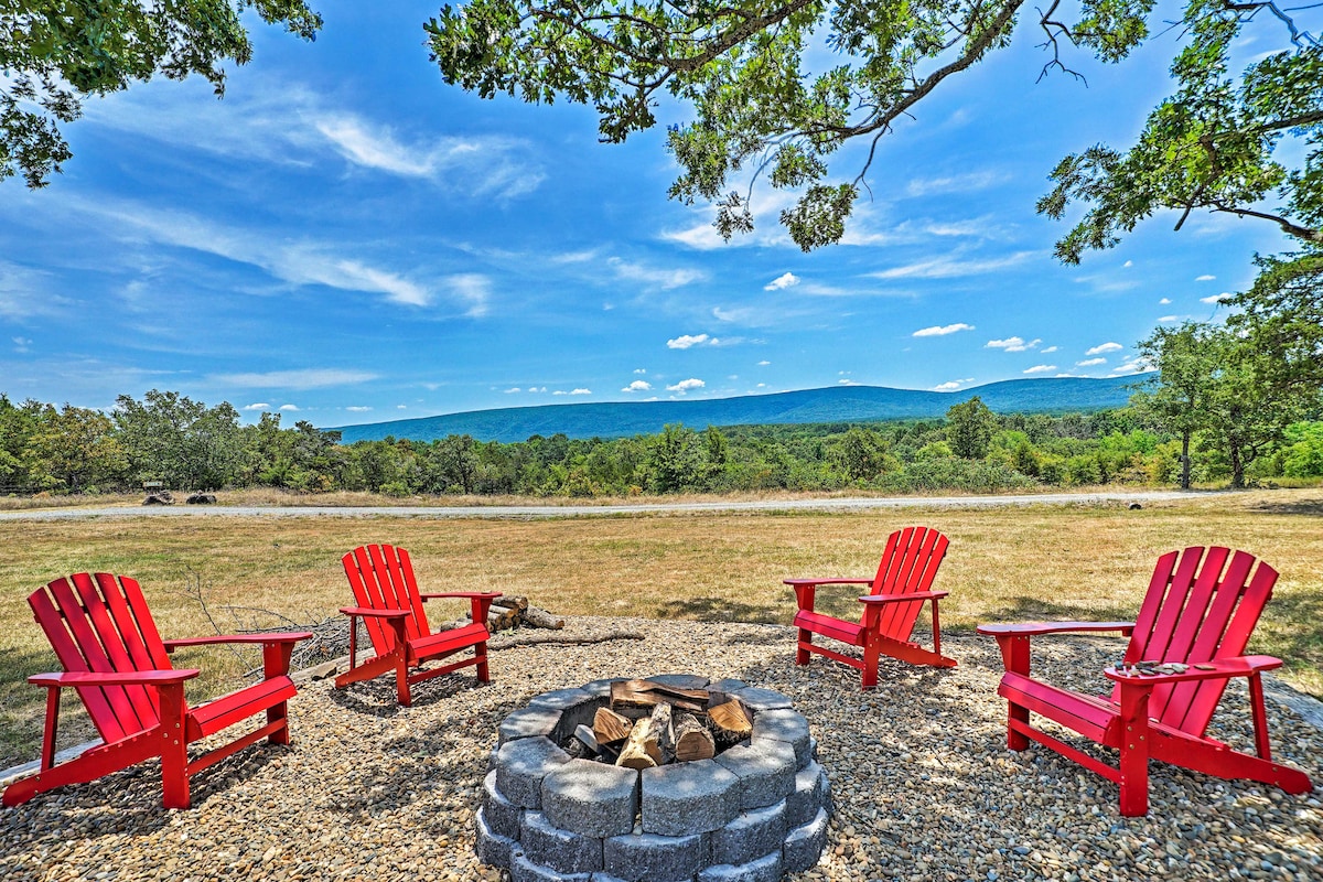 A fire pit constructed of grey stones is surrounded by four red Adirondack chairs, set against a backdrop of rolling green hills under a bright blue sky. The area features a gravel surface and is bordered by trees, enhancing the natural ambiance.