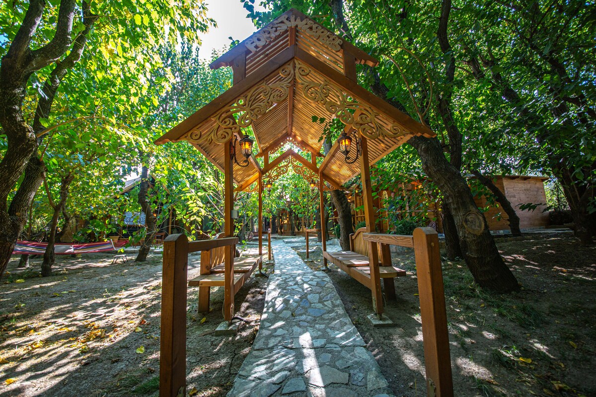 An inviting pathway made of stone leads through a verdant garden shaded by trees. A wooden structure with ornate detailing creates a charming gathering space, featuring benches under a sloped roof, illuminated by decorative lanterns that hang from the ceiling.
