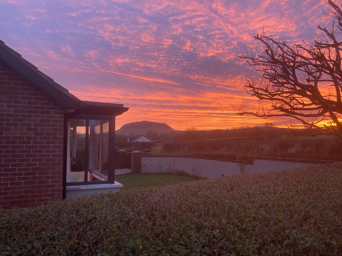 A vibrant sunset sky casts warm hues over the landscape, with Slemish mountain silhouetted in the distance. The image captures the cottage's exterior, framed by a hedge and the soft glow of glass windows reflecting the evening light.