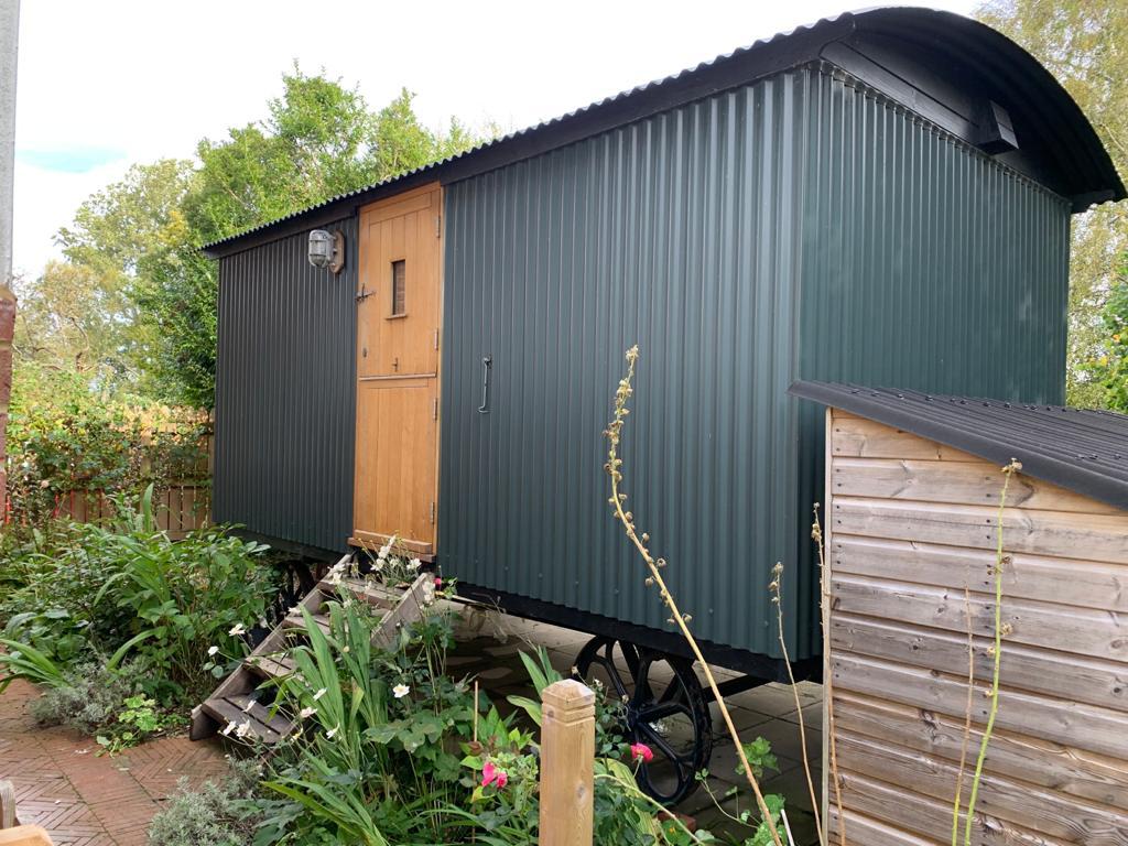 The shepherd's hut, featuring a stylish corrugated exterior, is nestled in a garden surrounded by greenery. Steps lead to the entrance, where a wooden door and small window are visible. The rustic charm of the hut is highlighted by the natural landscaping.