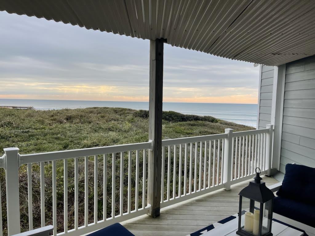 A private balcony offers a view of the ocean and dunes, framed by a white railing. A cozy seating area with navy cushions is positioned to enjoy the serene coastal landscape, while a lantern adds a touch of warmth to the inviting space.