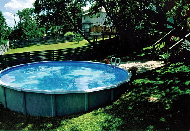 A round above-ground pool is surrounded by a grassy area, featuring clear blue water. Nearby, a wooden deck can be seen in the background along with a curving slide descending from a higher level. A few chairs are positioned beside the pool.