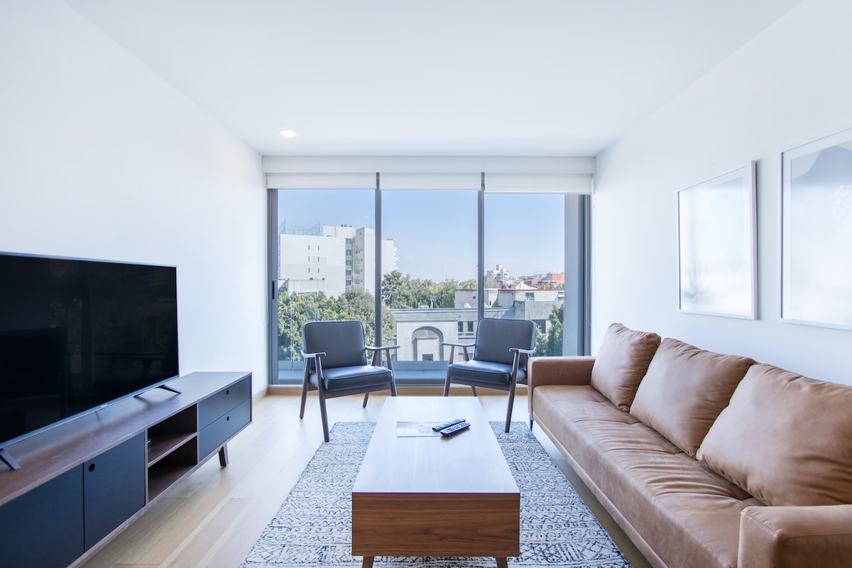 A modern living area is observed, featuring a brown sofa and two black armchairs positioned around a wooden coffee table. A flat-screen TV is mounted on a media console. Large windows allow ample natural light and offer a view of the cityscape.