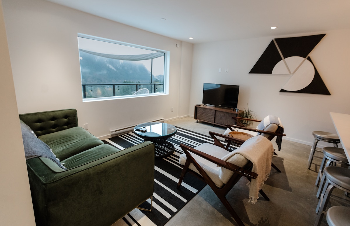 A contemporary living area features a green couch paired with wooden chairs, a round black coffee table, and a flat-screen television. Large windows offer mountain views, while geometric artwork enhances the neutral-toned space. A striped rug adds texture to the polished concrete floor.