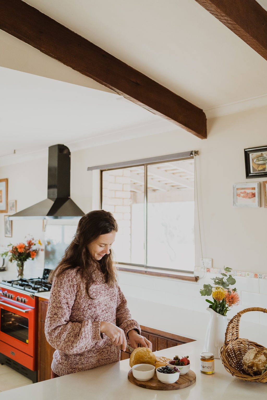 A bright kitchen features wooden beam ceilings and a large window. A person is seen preparing food on a countertop, with a basket and bowls of ingredients nearby. An inviting red stove adds a pop of color to the space, complemented by floral decor.