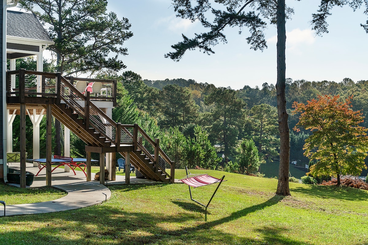 A grassy outdoor area is displayed, featuring a colorful hammock suspended between two trees. A wooden staircase leads to an upper deck, and a patio path meanders through the landscape. Lush trees and foliage provide a natural backdrop, with hints of the lake visible in the distance.