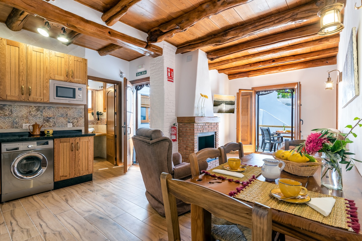 A rustic kitchen and dining area feature wooden cabinetry and a round dining table set with yellow dishes. A washing machine and microwave are positioned under a wooden shelf. Bright light flows in from open doors leading to an outdoor seating area.