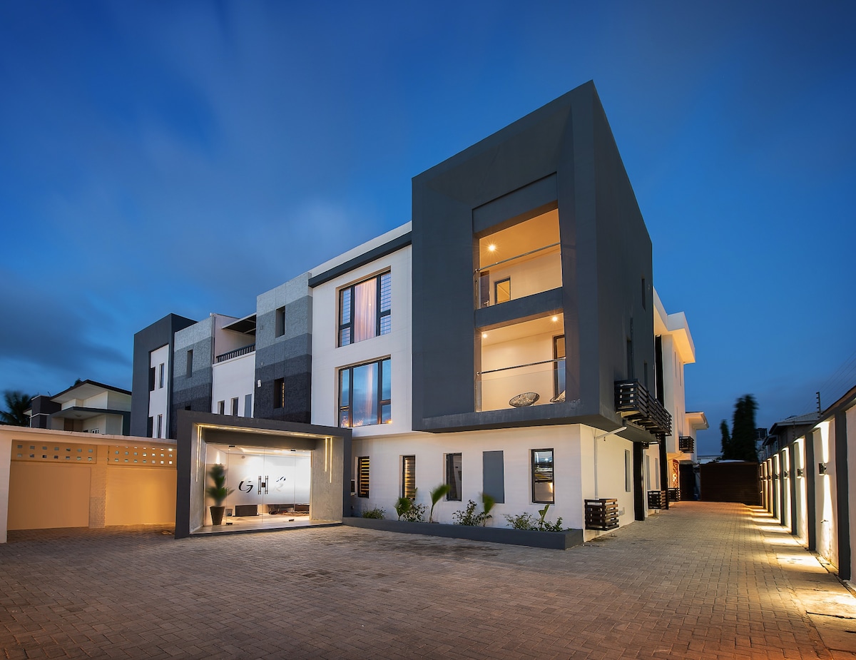 The exterior of the modern apartment building is illuminated at dusk, showcasing a sleek, contemporary design with a mix of dark and light materials. A spacious parking area is visible in the foreground, flanked by well-maintained landscaping.