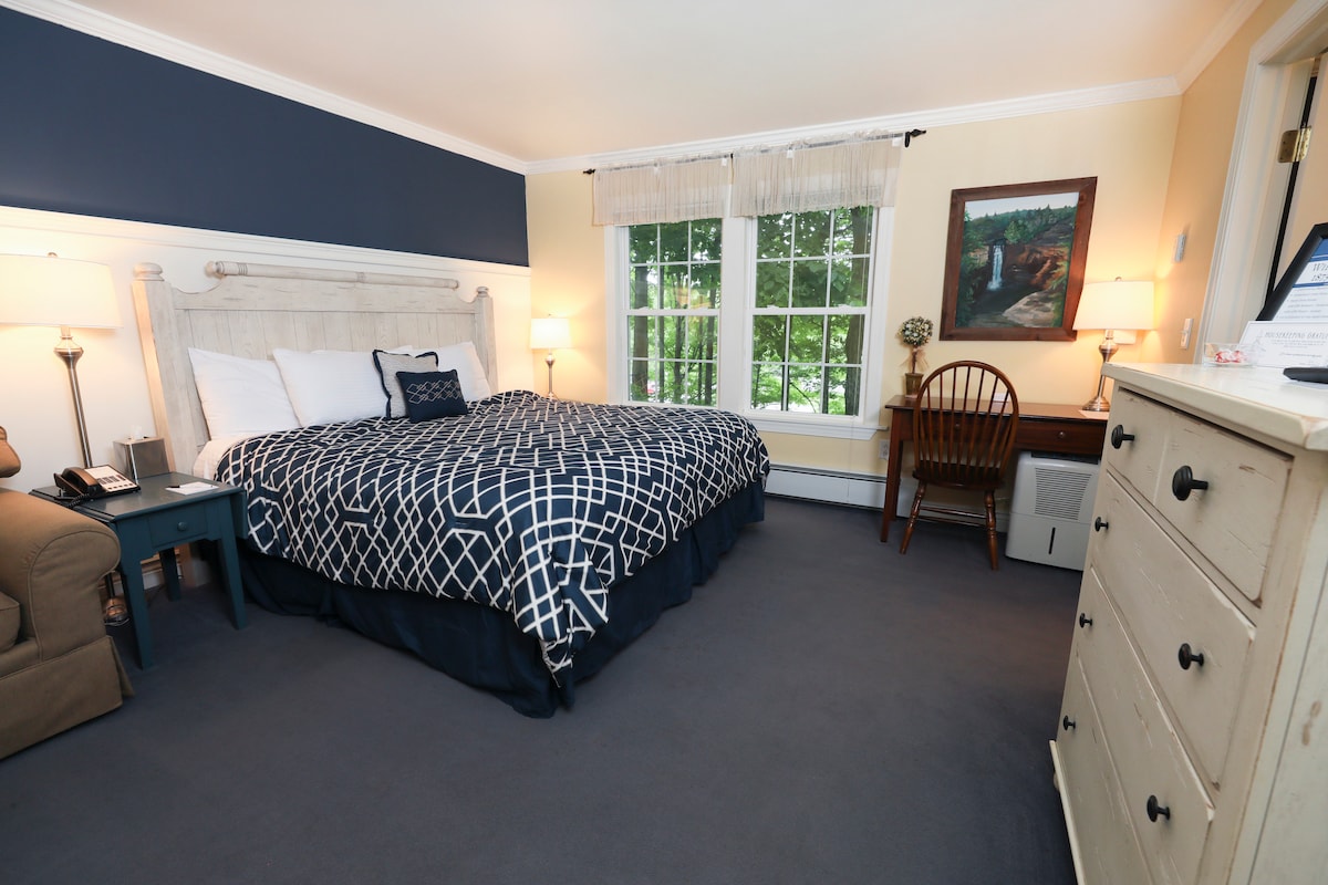 A spacious guest room features a plush king-sized bed with a navy and white patterned bedspread. A wooden desk and chair are positioned near the window, which is dressed with light curtains. A dresser stands opposite the bed, and a comfortable armchair rounds out the seating area.