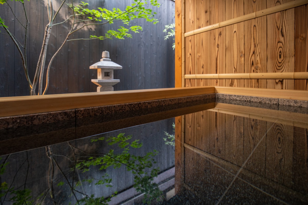 An outdoor hot spring bath reflects the wooden walls and surrounding stone lantern, creating a sense of tranquility. The smooth surface of the water mirrors the natural elements, while a tree adds a touch of greenery to the serene atmosphere.