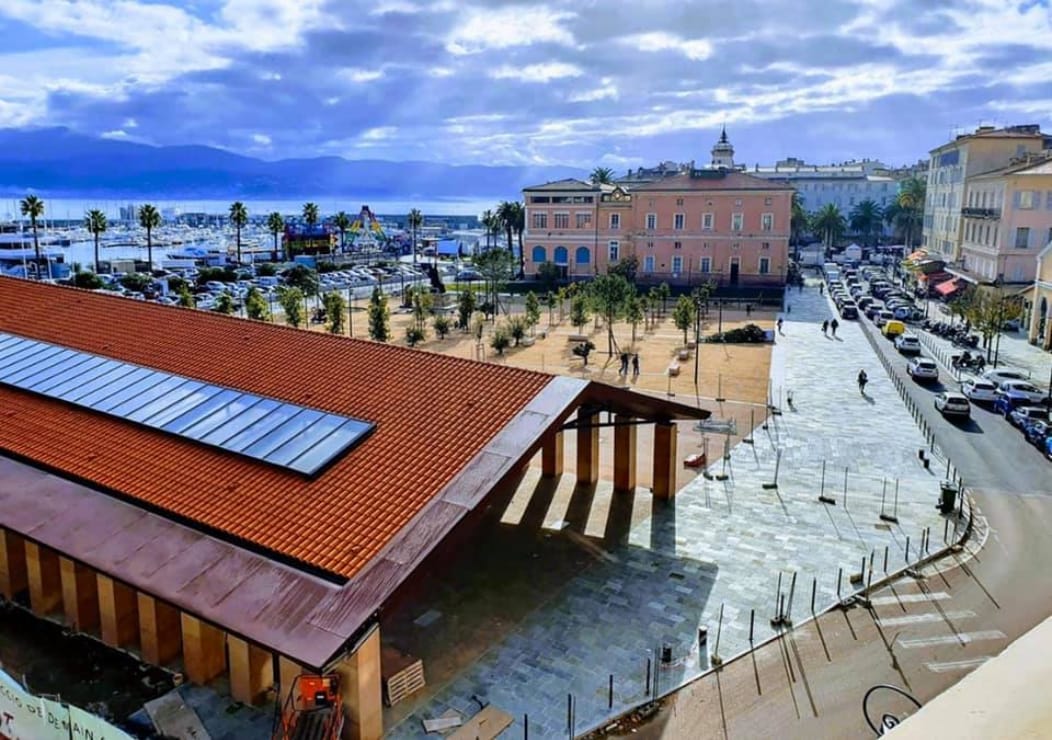 An outdoor view captures a bustling square with a modern structure featuring a wooden roof. Surrounding palm trees and open green areas are visible, alongside a distant glimpse of the waterfront. Streets lined with vehicles and buildings add to the urban landscape.