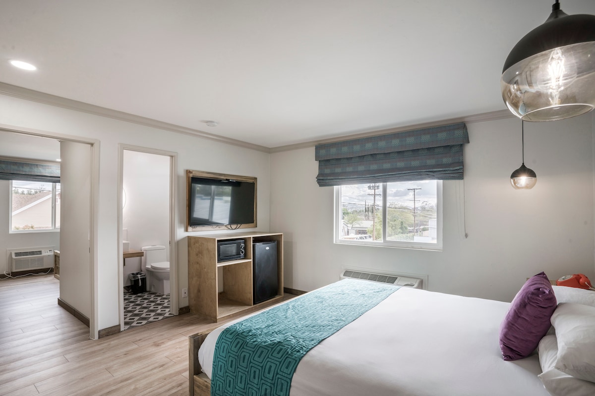 A well-lit room features a queen-sized bed with an aqua-colored throw and purple accent pillows. A flat-screen TV and a mini-fridge are positioned at the foot of the bed. A modern bathroom is visible through a partially opened door, with patterned tiles and natural light streaming through the window.