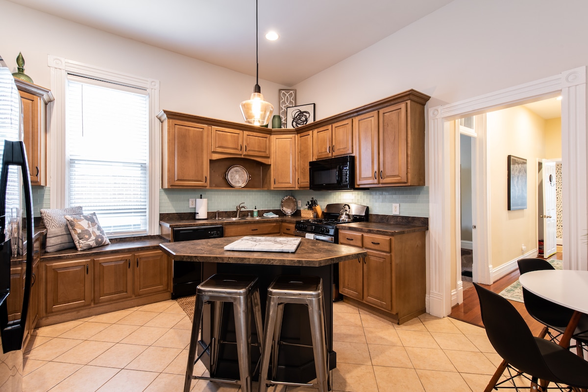 The kitchen features warm wooden cabinetry and modern appliances, including a black refrigerator and microwave. A central island provides additional prep space, complemented by metal stools. Natural light fills the room through the windows, enhancing the spacious layout, while a doorway leads to another area of the home.