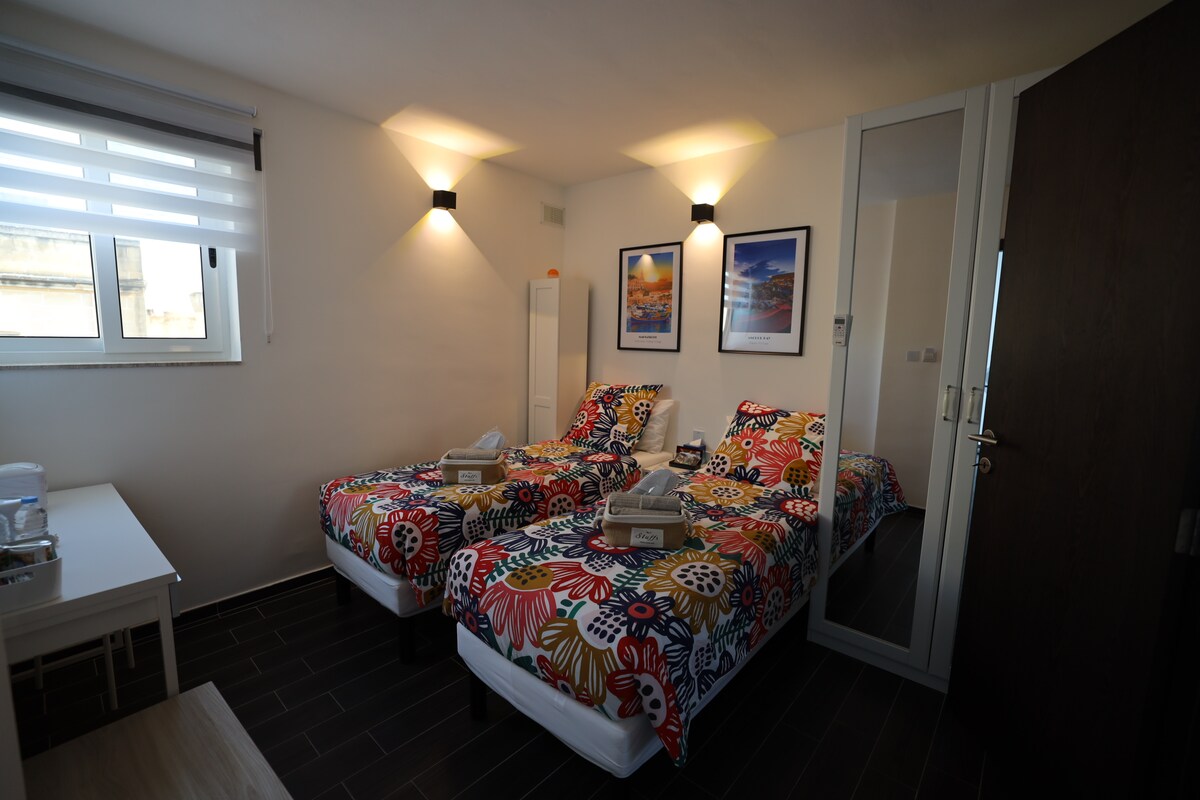 The room features two twin beds adorned with vibrant floral patterns. A small table is positioned beside the beds, and a tall mirror stands against the wall. Light is provided by wall sconces, and natural light enters through a window dressed with layered shades.