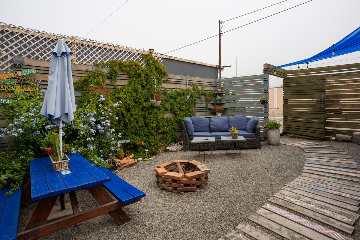 An outdoor patio features a blue wooden picnic table and a comfortable sofa set, surrounded by greenery and flowers. A fire pit sits at the center, while a large umbrella provides shade. The area is lined with wooden planks, creating a relaxed, cozy atmosphere.