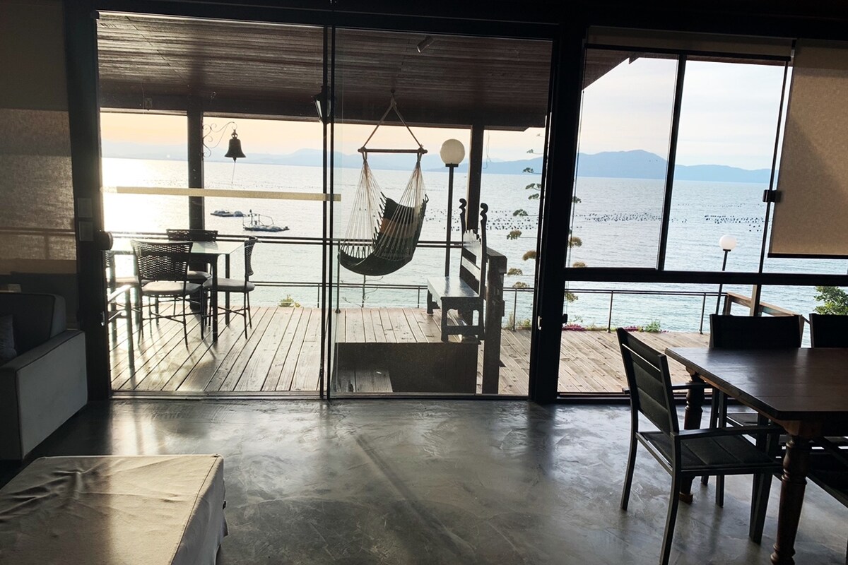 A spacious open-plan area features large glass doors providing a view of the ocean. A hammock is suspended from the ceiling, and a dining table is positioned to the side. The natural light enhances the neutral-toned interior, while the deck outside leads to the tranquil beach.