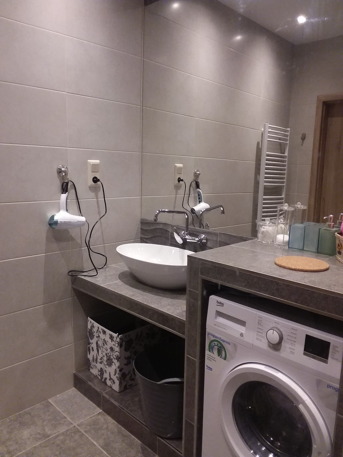 The bathroom features modern tiled walls and flooring, with a round sink set into a countertop. A washing machine is positioned at the bottom right. Two faucets and hairdryer are mounted on the wall beside the sink. A towel rack and various toiletries can be seen.