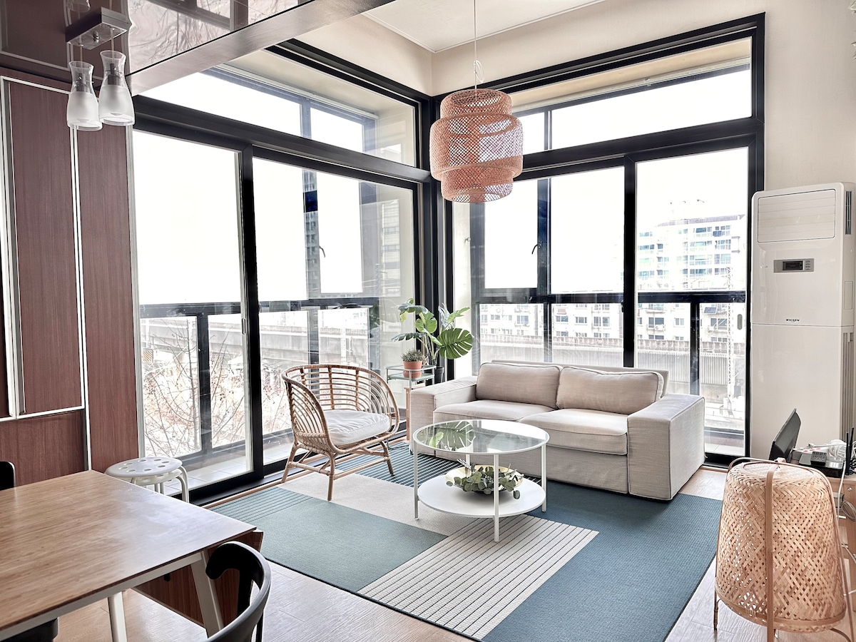 A bright living area is defined by large windows that provide ample natural light. A comfortable sofa sits alongside a circular coffee table, with a plant adding a touch of greenery. A dining area is visible, featuring wooden furniture and a woven light fixture.