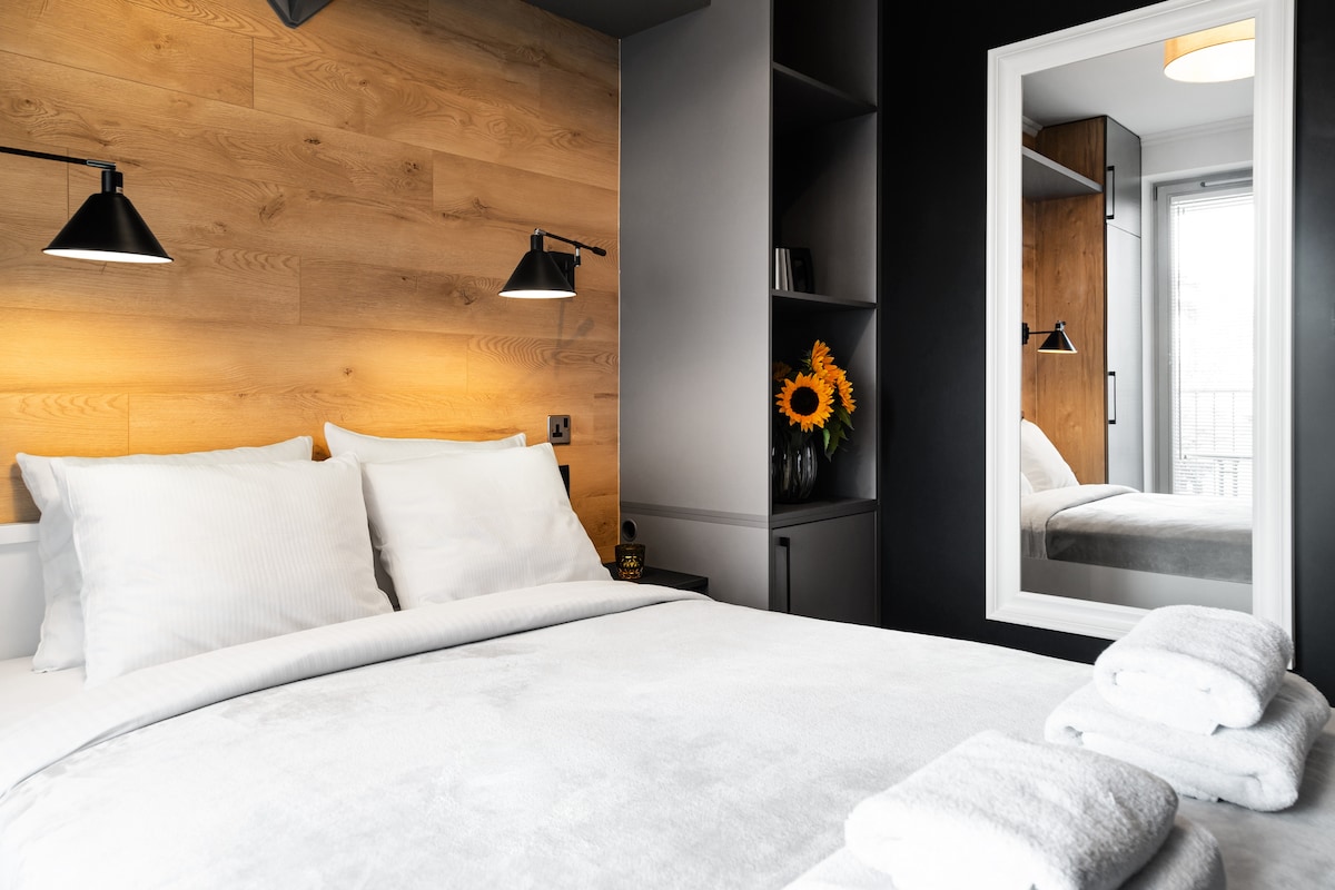 A bedroom is featured with a spacious bed adorned with neutral bedding and neatly arranged towels. A warm wooden accent wall complements modern shelving, while a mirror reflects the room's calm atmosphere. Sunflowers add a touch of freshness near a softly lit bedside.