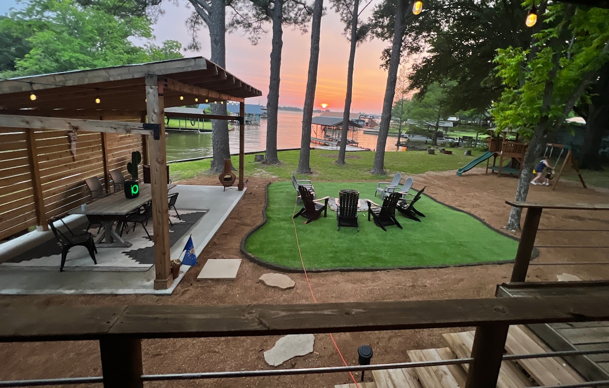 A lakeside area features a spacious turf fire pit surrounded by multiple adirondack chairs. The scene is complemented by a covered pavilion with a dining area, with picturesque sunset views visible over the water through towering pine trees.