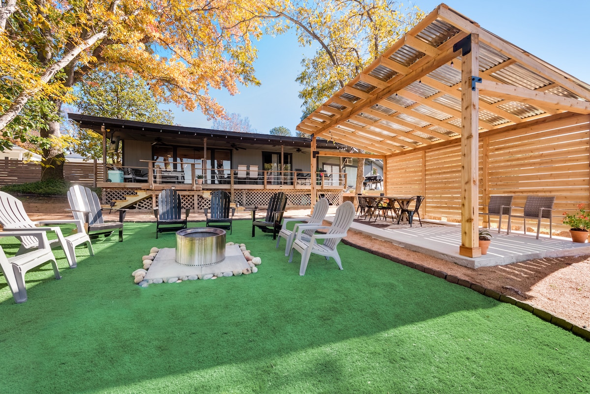 An outdoor space features a spacious green turf area with a fire pit surrounded by several seating options. A cedar pavilion provides shade, while a large deck and dining area are visible in the background, offering an inviting relaxation space.