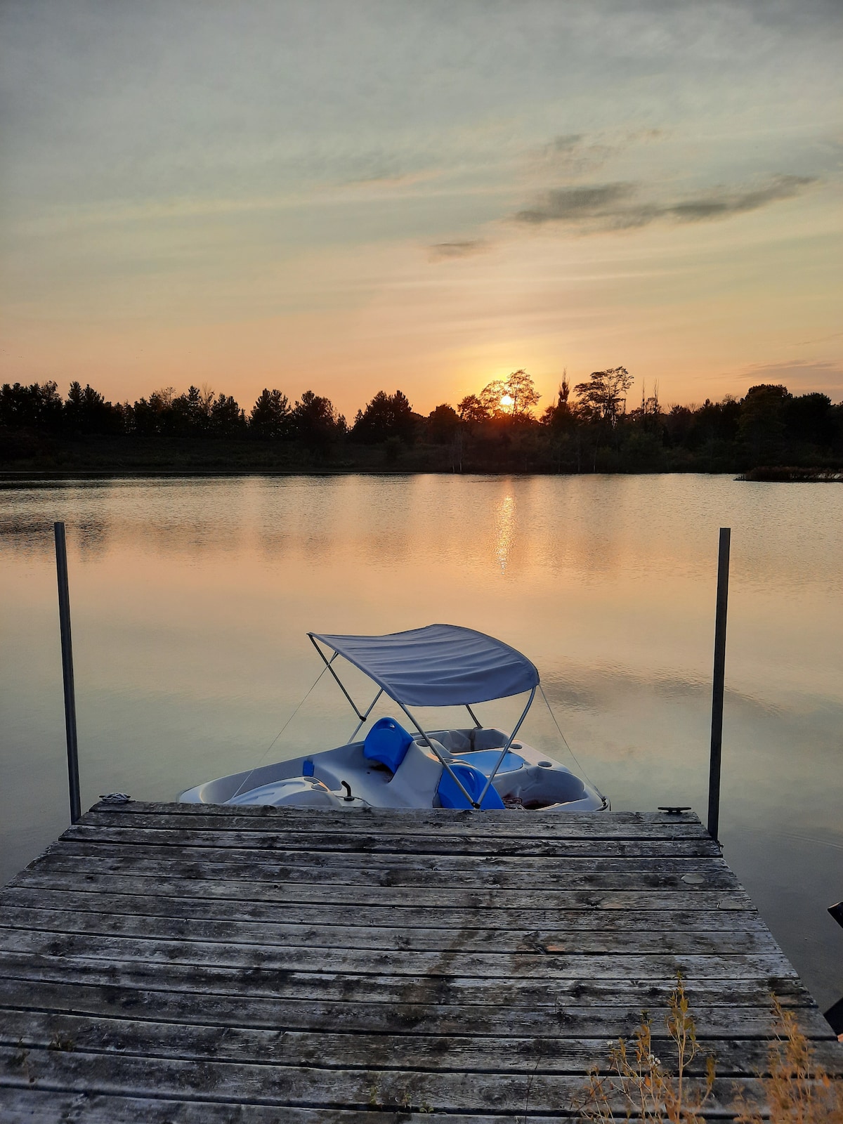 A wooden dock extends over calm waters, where a pedal boat rests gently at the edge. The sun sets in the background, casting warm hues across the lake's surface. Silhouettes of trees line the opposite shore, creating a serene natural setting.