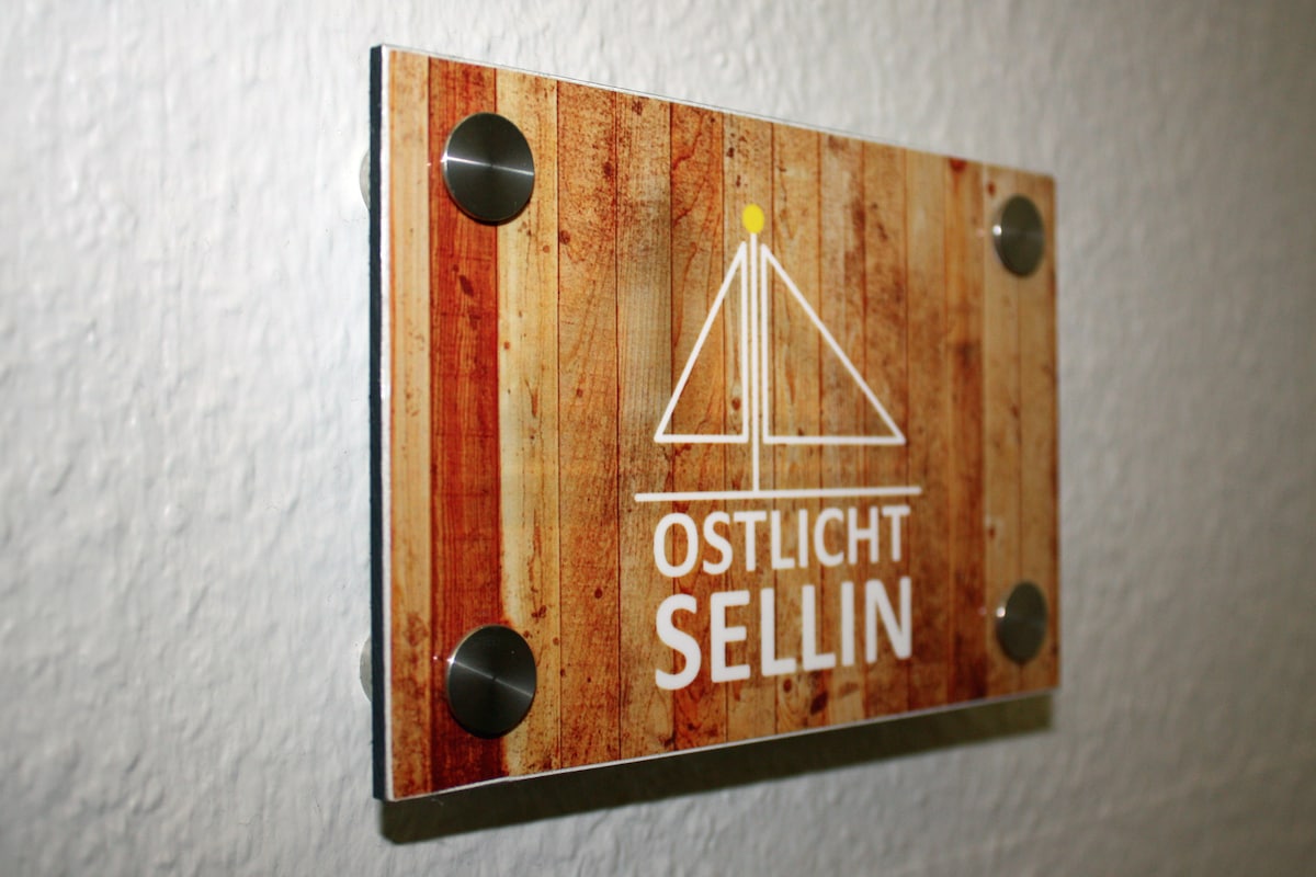 A wooden sign displaying the name 'Ostlicht Sellin' is affixed to a textured wall. The sign features a lighthouse graphic above the name, with a sun symbol positioned above it, adding a nautical theme to the design.