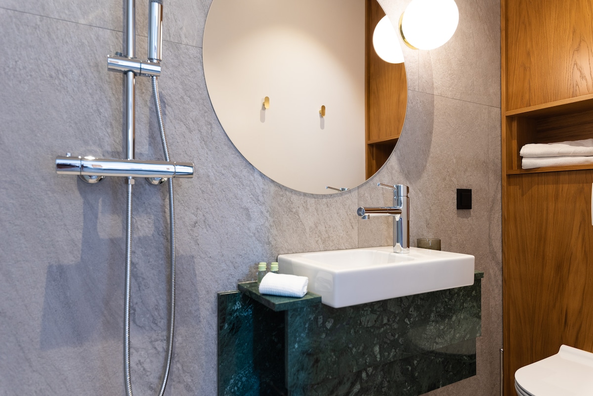 A modern bathroom features a sleek sink set atop a green marble countertop. A round mirror is mounted above the sink, and contemporary lighting fixtures are installed on the wall. Shower fixtures are positioned beside the sink, and wood shelves hold neatly arranged towels.