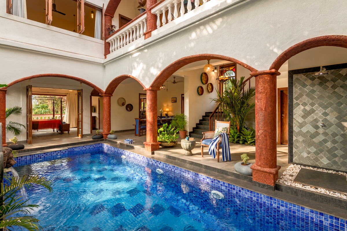 An open-air courtyard features a vibrant blue swimming pool surrounded by arched support columns. Lush greenery adds a natural touch, while decorative elements are visible on the walls. Indoor dining and seating areas can be seen in the background, enhancing the inviting atmosphere.