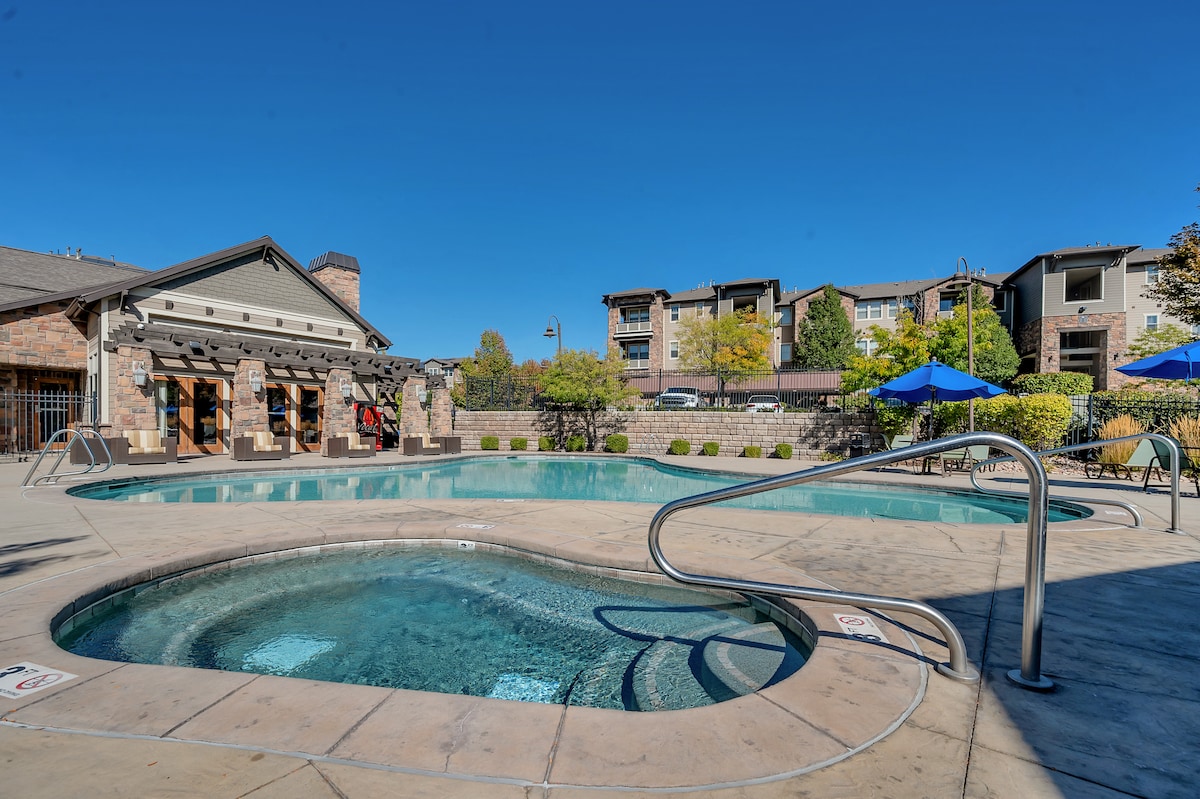 A large outdoor space features a swimming pool surrounded by blue lounge chairs and shaded umbrellas. A nearby hot tub is positioned at the pool's edge. Stone walls and greenery create a welcoming atmosphere, with a clear blue sky overhead.