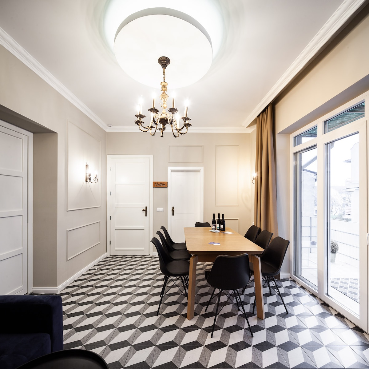 A dining area features a large wooden table surrounded by black chairs. A stylish chandelier hangs from the ceiling, and light enters through wide glass doors leading to an outdoor space. The geometric floor design adds a modern touch to the room.
