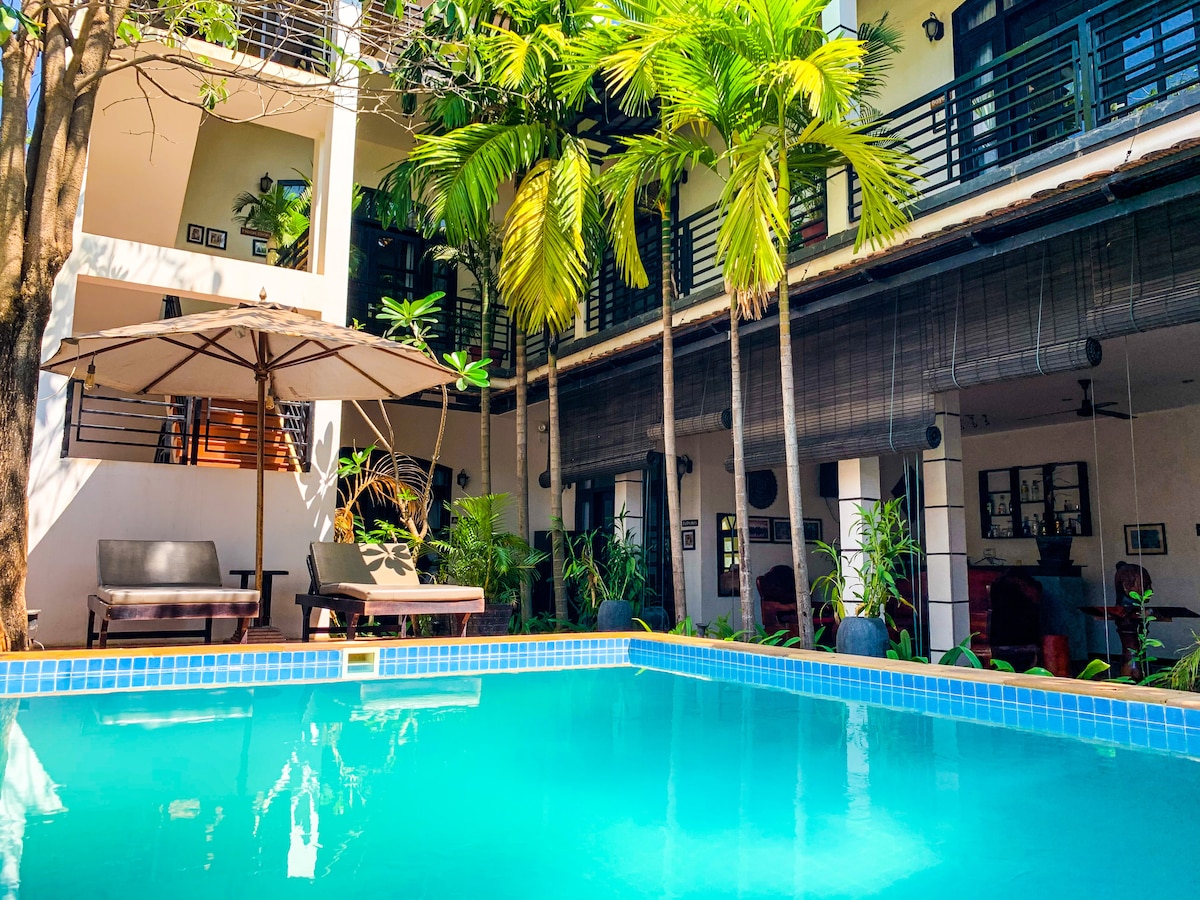 A private pool features clear turquoise water, surrounded by lush palm trees and greenery. Comfortable lounge chairs are positioned under a large umbrella, offering a serene space for relaxation. The pool area is complemented by a relaxed ambiance created by the open layout of the surrounding architecture.