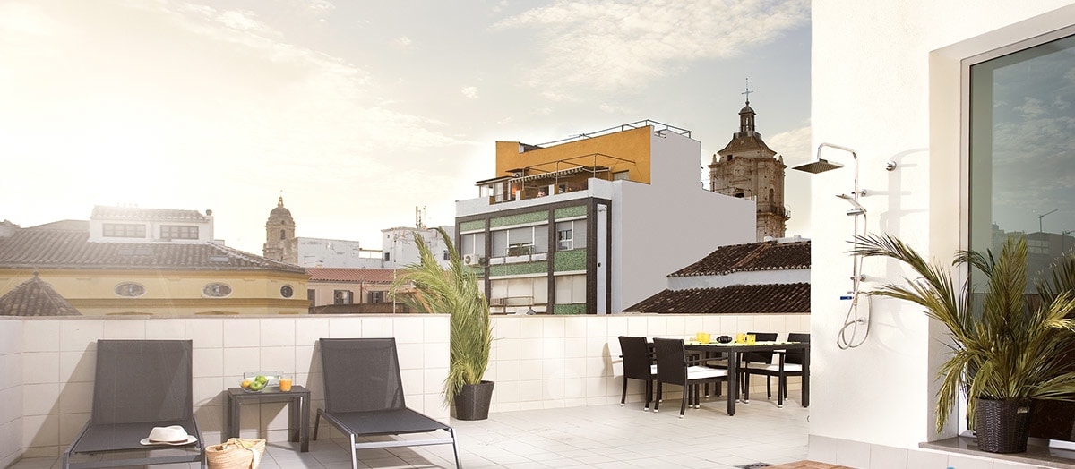 A shared terrace with outdoor seating is visible, featuring a table with chairs alongside lounge chairs. The view captures nearby buildings under a bright sky, highlighting a mix of architectural styles. Potted plants add greenery to the space, creating an inviting atmosphere.