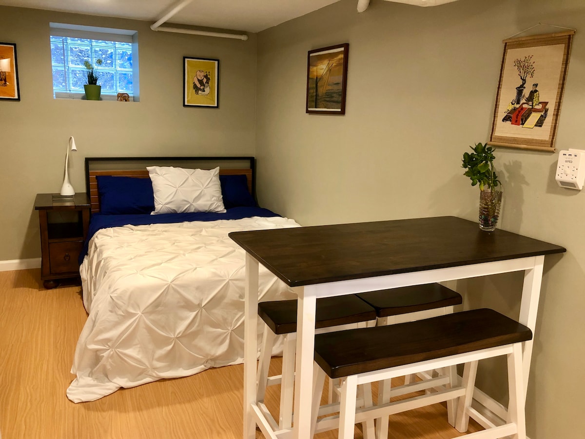 A cozy studio space features a queen size bed with a light-colored blanket against a green wall. A snug bar table and stools are nearby, with framed art adorning the walls. Natural light enters through a window above the bed, adding warmth to the atmosphere.