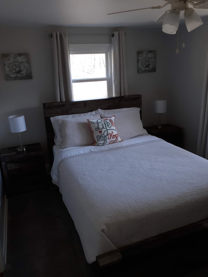 سرير كوين 1 مع طاولات جانبية للسرير وخزانة ملابس مع مرآة مع علّاقات ومنافذ في المصابيح.  توجد مروحة سقف/مشط إضاءة ونوافذ على جانبي الغرفة.