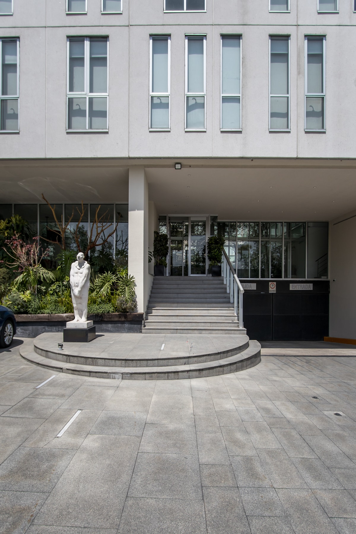 The entrance of a modern building is highlighted by a curved stone staircase leading to a glass door. A contemporary statue stands on a round pedestal at the base of the stairs. Lush green landscaping is visible alongside the steps.