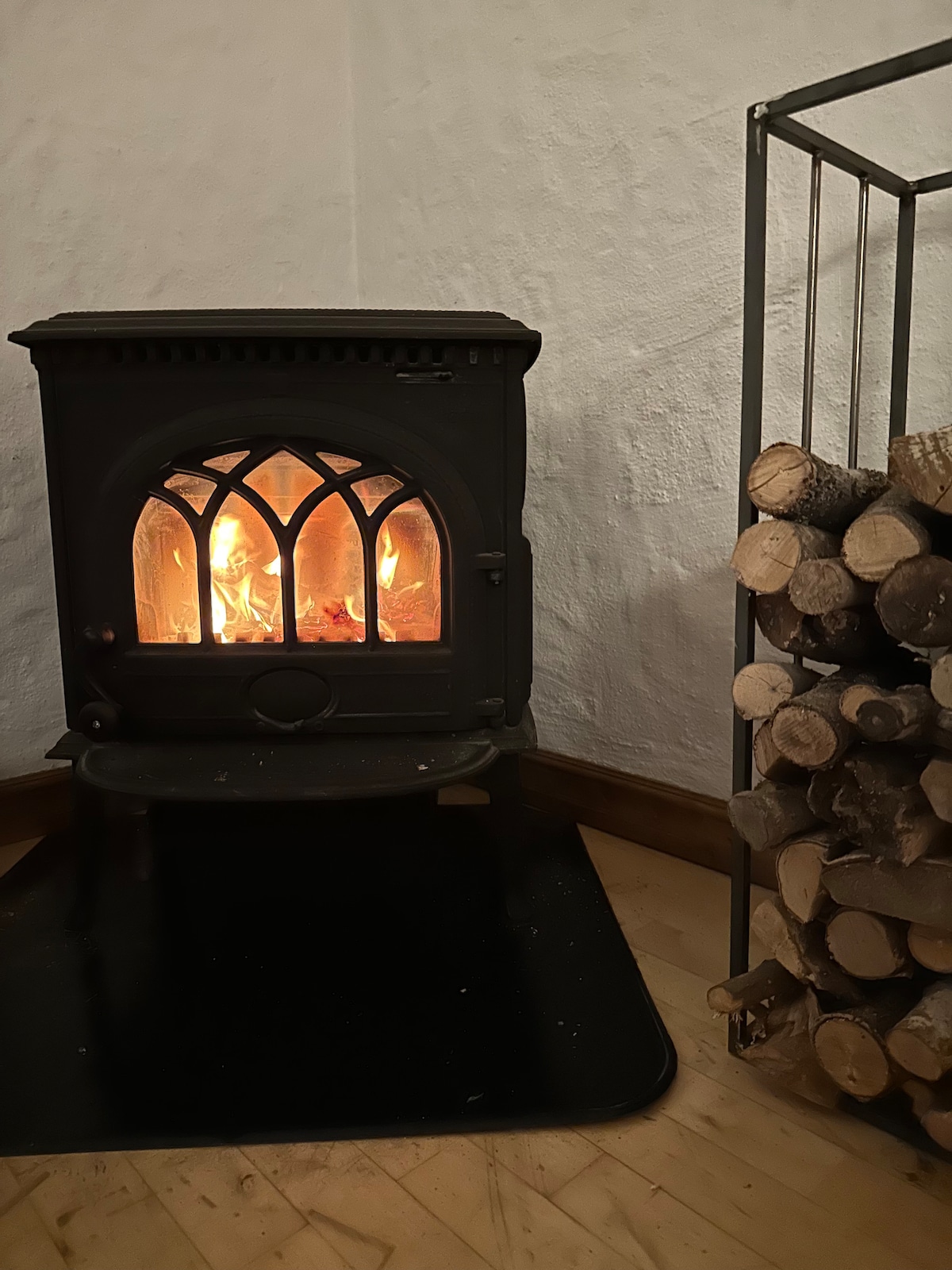 A wood-burning stove is positioned prominently against a textured white wall, its glowing flames visible through an arched window design. A metal wood holder nearby displays neatly stacked logs, contributing to a cozy, functional atmosphere.