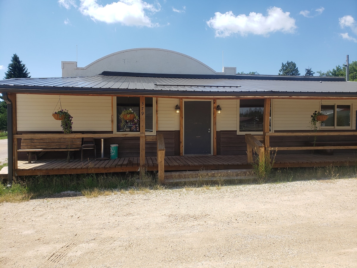 The exterior of the building features a welcoming porch with wooden seating and flower planters. A gravel path leads to the entrance, which is framed by large windows. The roof is metallic, and the structure has a classic architectural design.