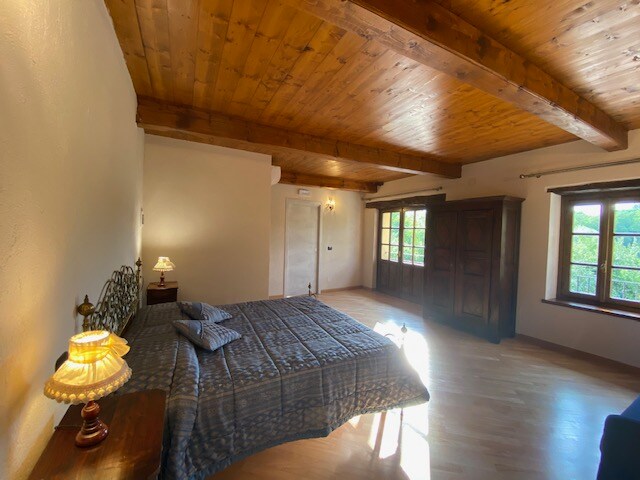 An elegant room features a spacious bed dressed in a subtle blue coverlet. Exposed wooden beams enhance the ceiling. Natural light enters through several windows, illuminating the warm wooden floor. A wooden wardrobe and a bedside lamp add to the room's charm.