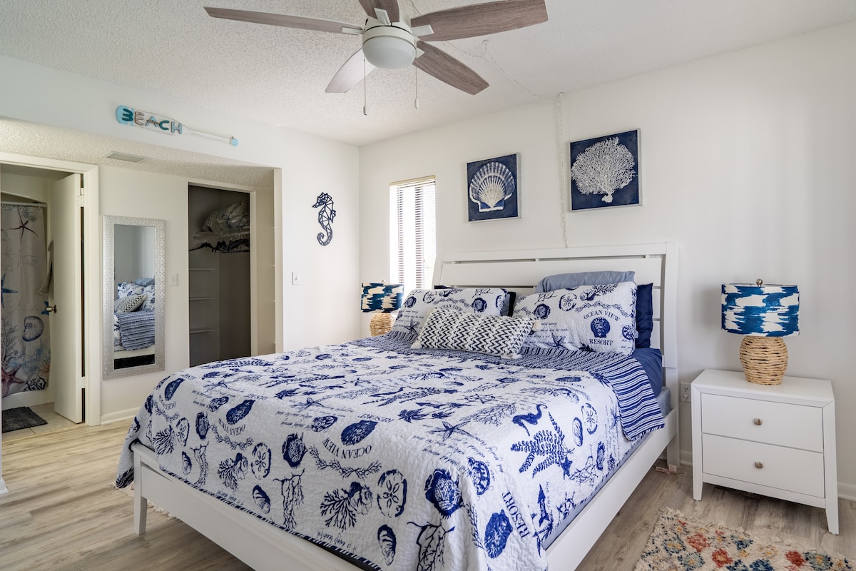 A bright bedroom features a spacious white bed adorned with a blue and white patterned quilt. Two decorative lamps with woven bases flank the bed. Soft light enters through a window covered by sheer curtains, enhancing the relaxed ambiance of the room.