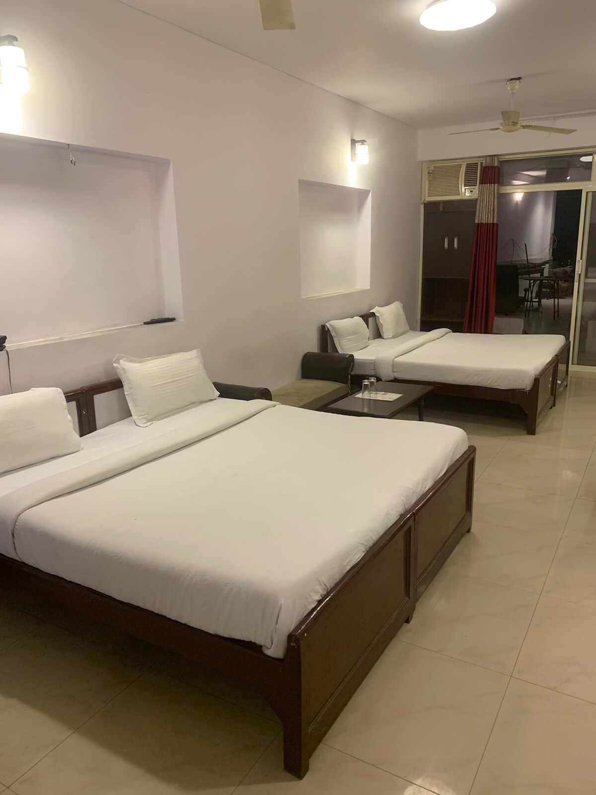 A spacious room is furnished with two beds, complemented by simple wooden frames and crisp white linens. A small table is positioned between the beds, providing added convenience. Large windows allow natural light to fill the space, with a seating area visible in the corner.