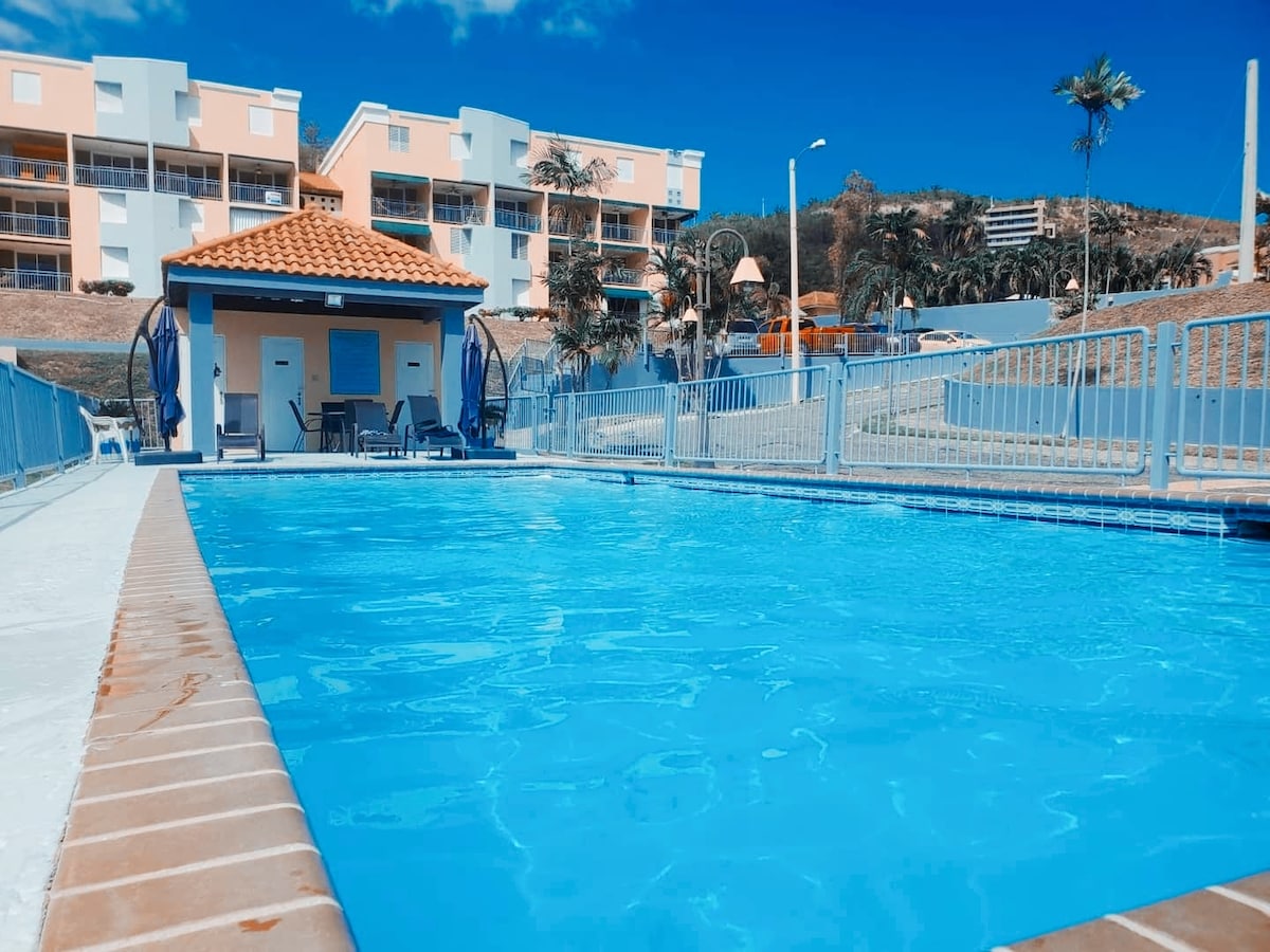 The swimming pool area is outlined by a secure fence and features clear, inviting water. Sun loungers are placed nearby, and a small clubhouse stands with a roof in the background, surrounded by palm trees and green landscape.
