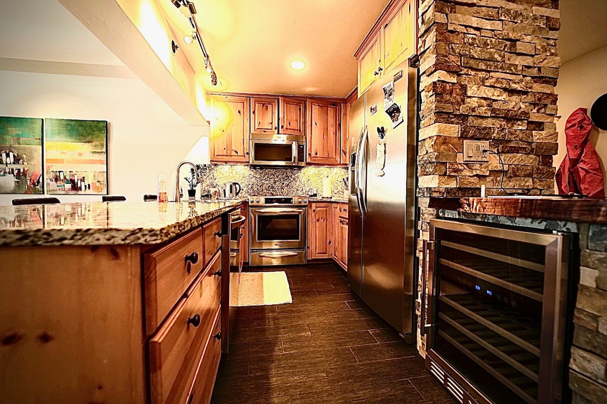 The kitchen features wooden cabinetry paired with granite countertops, offering ample workspace. Stainless steel appliances include an oven, microwave, and refrigerator. A wine cooler is positioned near the stone wall. Under-cabinet lighting enhances the workspace, and an inviting area rug adds warmth.