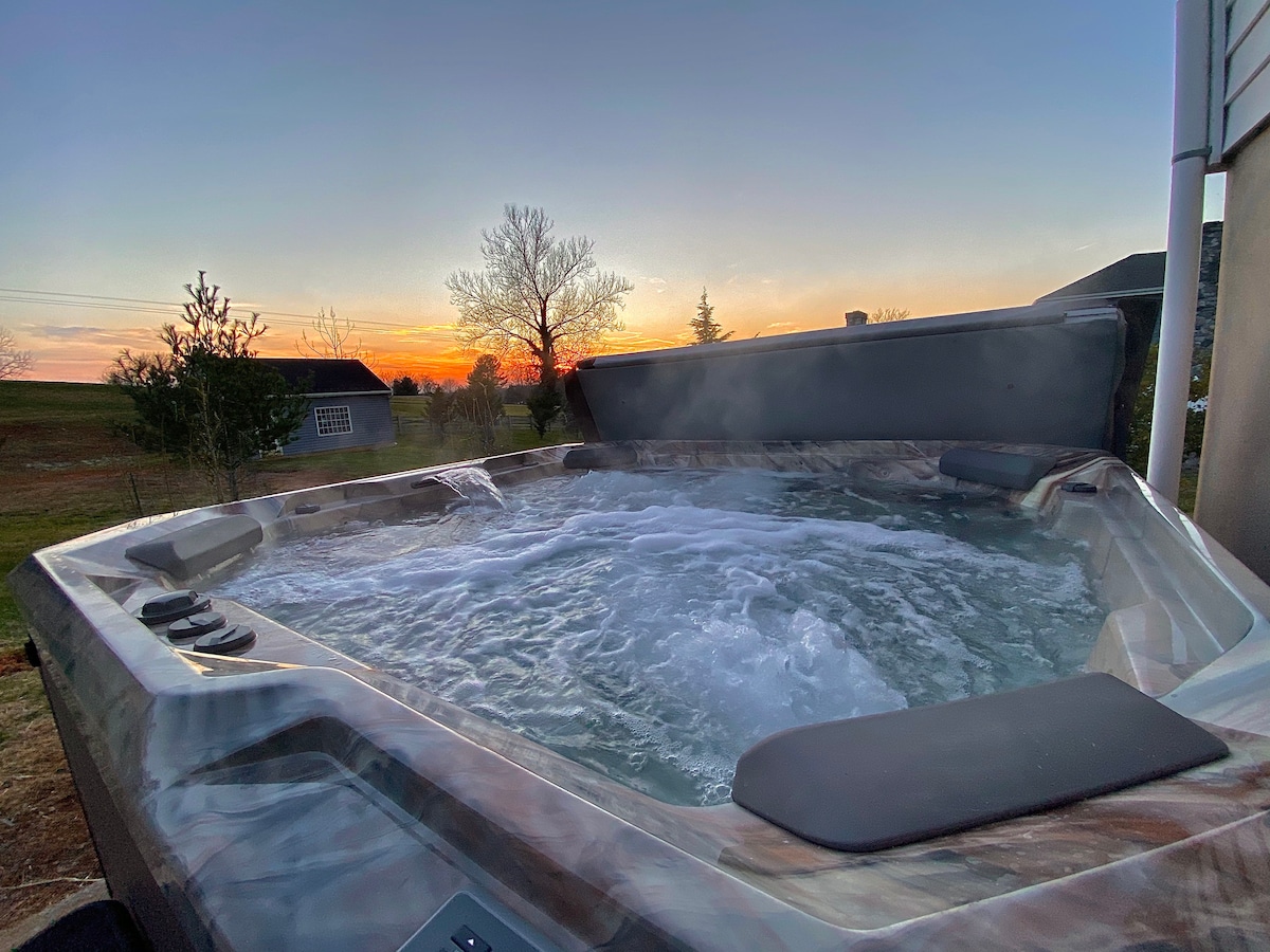 A hot tub is situated on a patio, steam rising gently in the cool evening air. The setting sun casts warm colors across the sky, providing a tranquil backdrop. Nearby, silhouettes of trees and buildings enhance the serene evening ambiance.